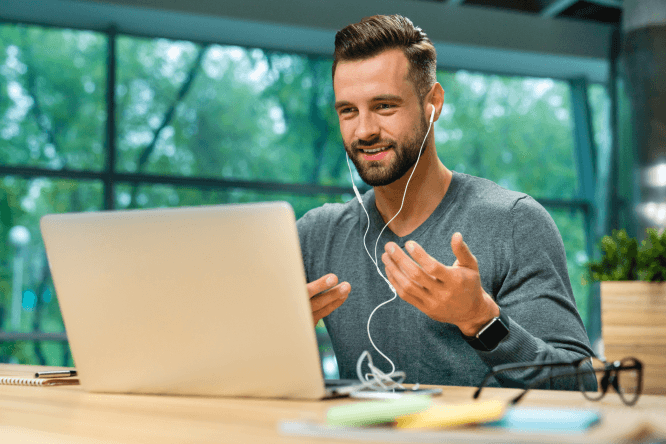 Young man wearing sweater is sitted in front of his laptop and having an online course while talking using his headset.