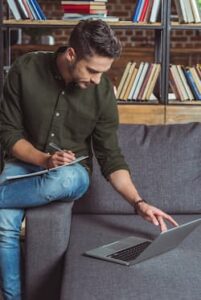 Handsome young man studying with notebook and laptop in library, wearing a pair of jeans and a khaki shirt