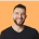 happy young man with brown hair and beard is smiling and wearing a black coloured t-shirt on orange background