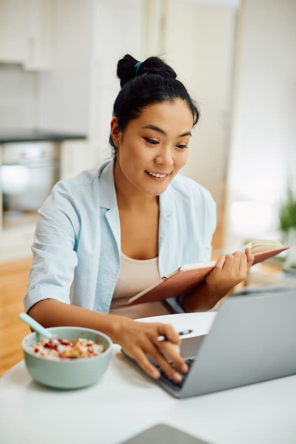 Smiling Asian woman using laptop and taking notes while having breakfast in the morning at home.