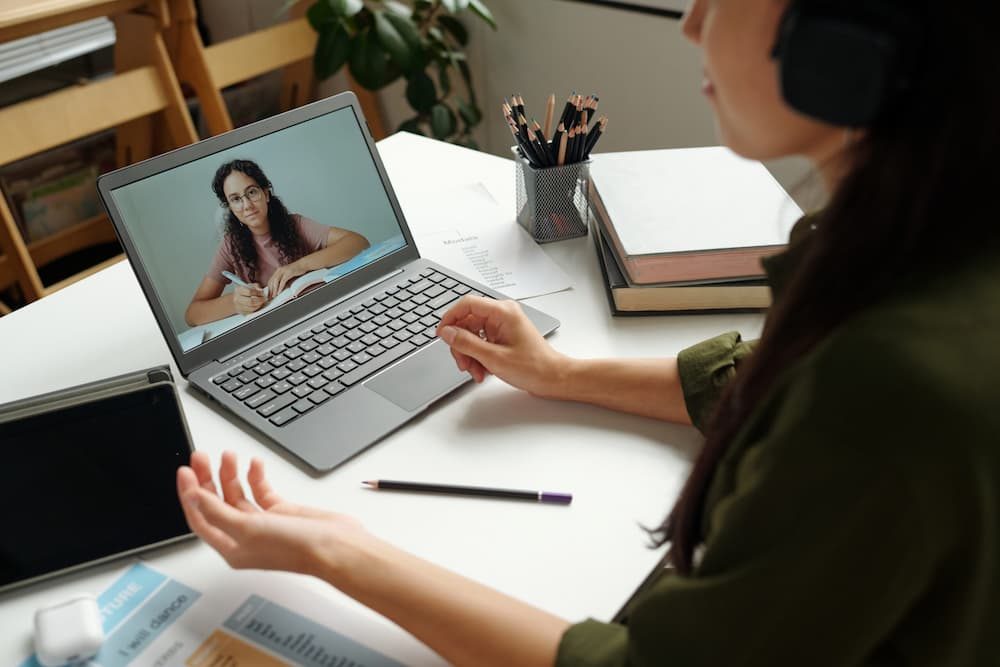 Focus on screen of laptop with clever schoolgirl making notes in copybook and listening to the explanation her female teacher is doing during the online lesson.