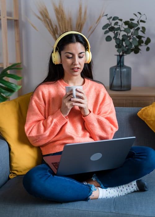Positive young female freelancer in casual clothes sitting on sofa and holding a mug, is browsing laptop while studying foreign languages remotely